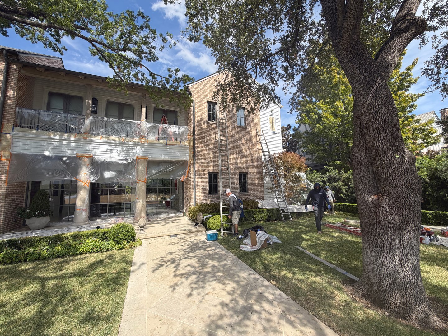 Wide view of a house with masked porch and windows, ladders, and a painting crew during exterior work.