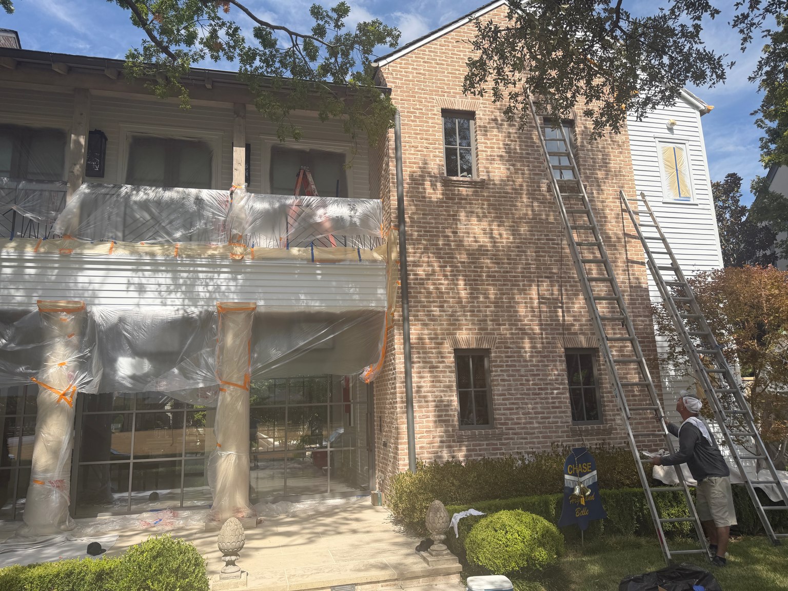 Closer view of a house exterior with windows and porch surfaces protected by plastic and tape during painting preparation.