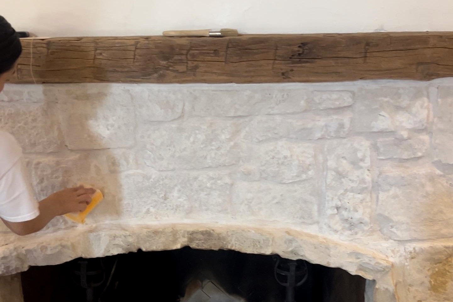Worker using a yellow sponge on the pale limewashed stone of a fireplace beneath a wood mantel.