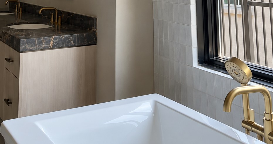 Bathroom with softly textured pale walls, a dark stone vanity, brass fixtures and a white tub.