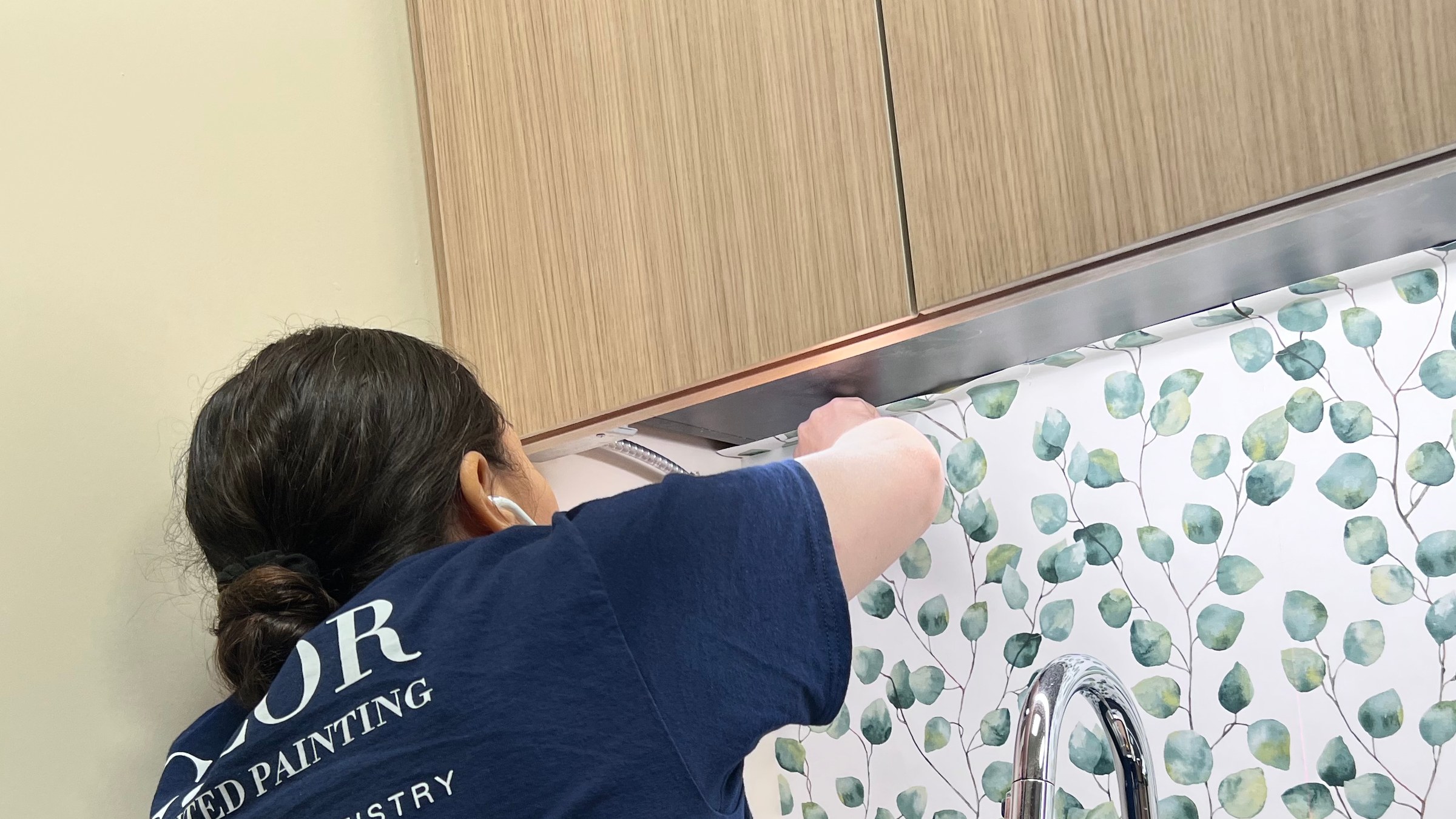 Worker positioning botanical-pattern wallpaper beneath kitchen cabinets beside a sink.