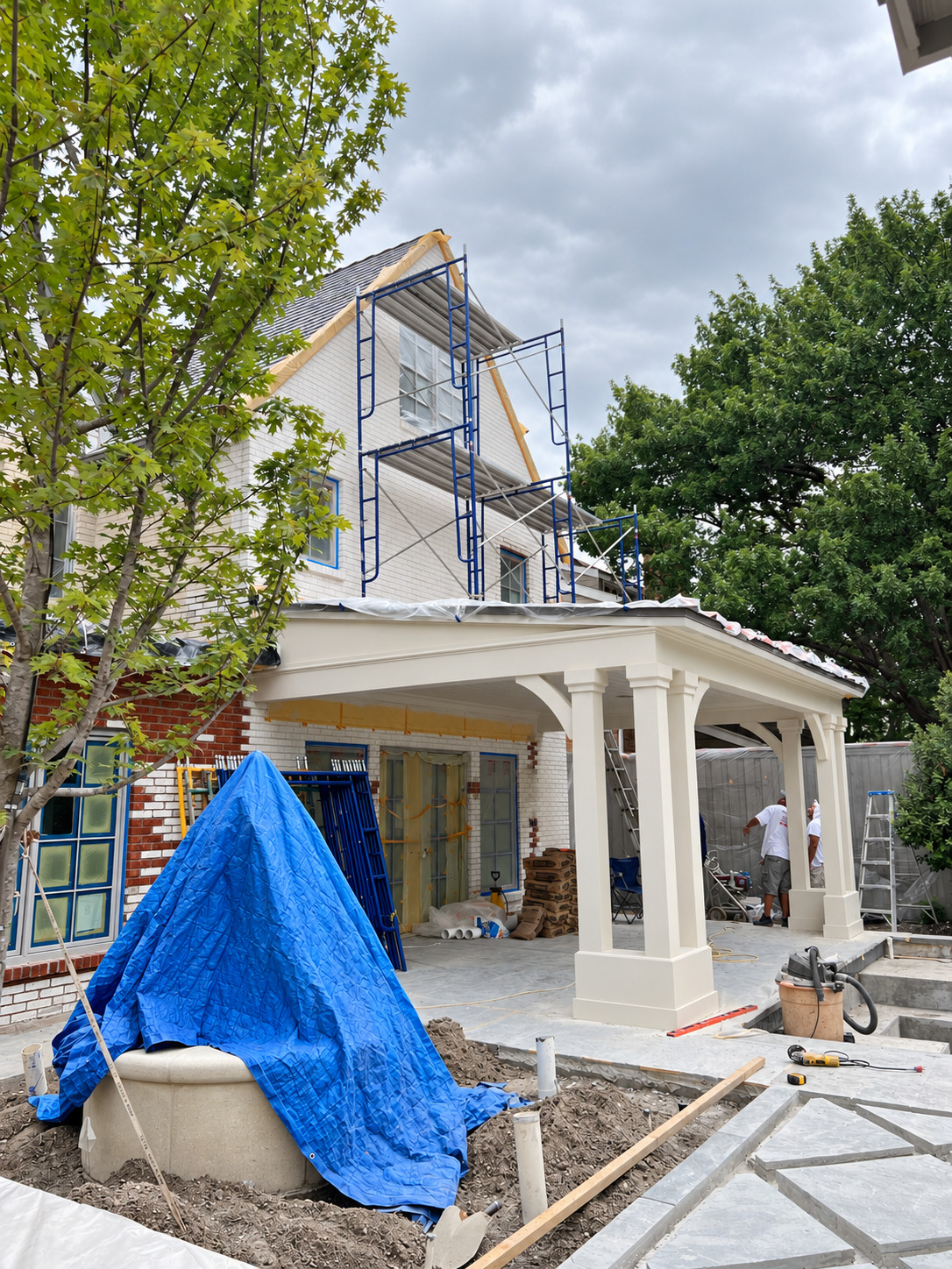 Two-story brick house during exterior work, with blue scaffolding, masked windows, white porch columns and protected work areas.