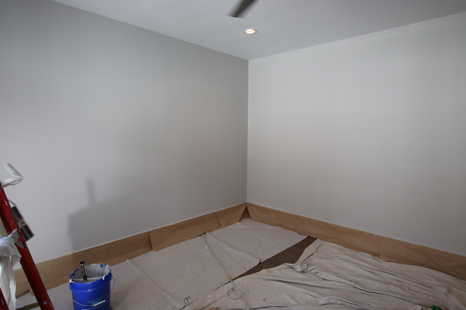 Smooth light walls above paper-masked baseboards, with drop cloths, a paint bucket and a ladder at the left edge.